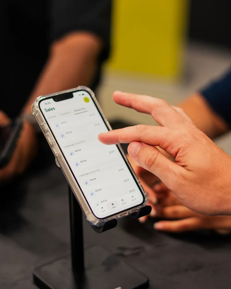 A phone mounted on a stand, showing the Teya Sales screen, a hand touching it